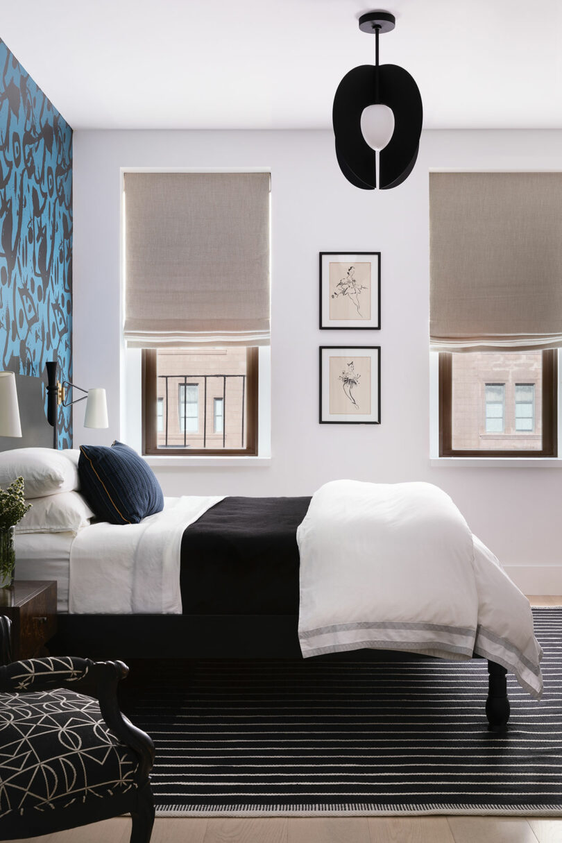 Modern bedroom with a single bed, black and white bedding, patterned accent wall, two beige tinted windows, three framed artworks, black chandelier and a striped rug.