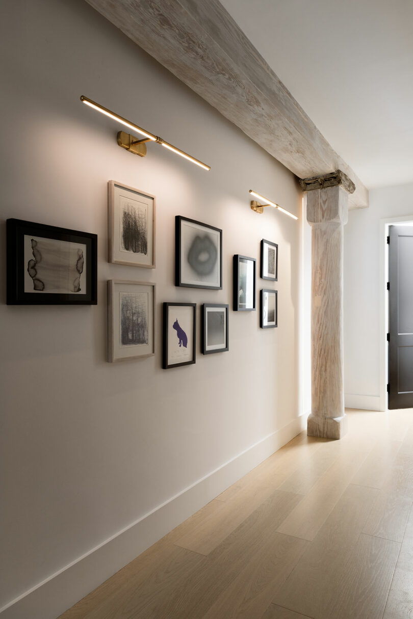 Hallway with framed artwork on wall, lit by two linear lights.  The space features light wood floors and an exposed wood beam next to an open doorway.