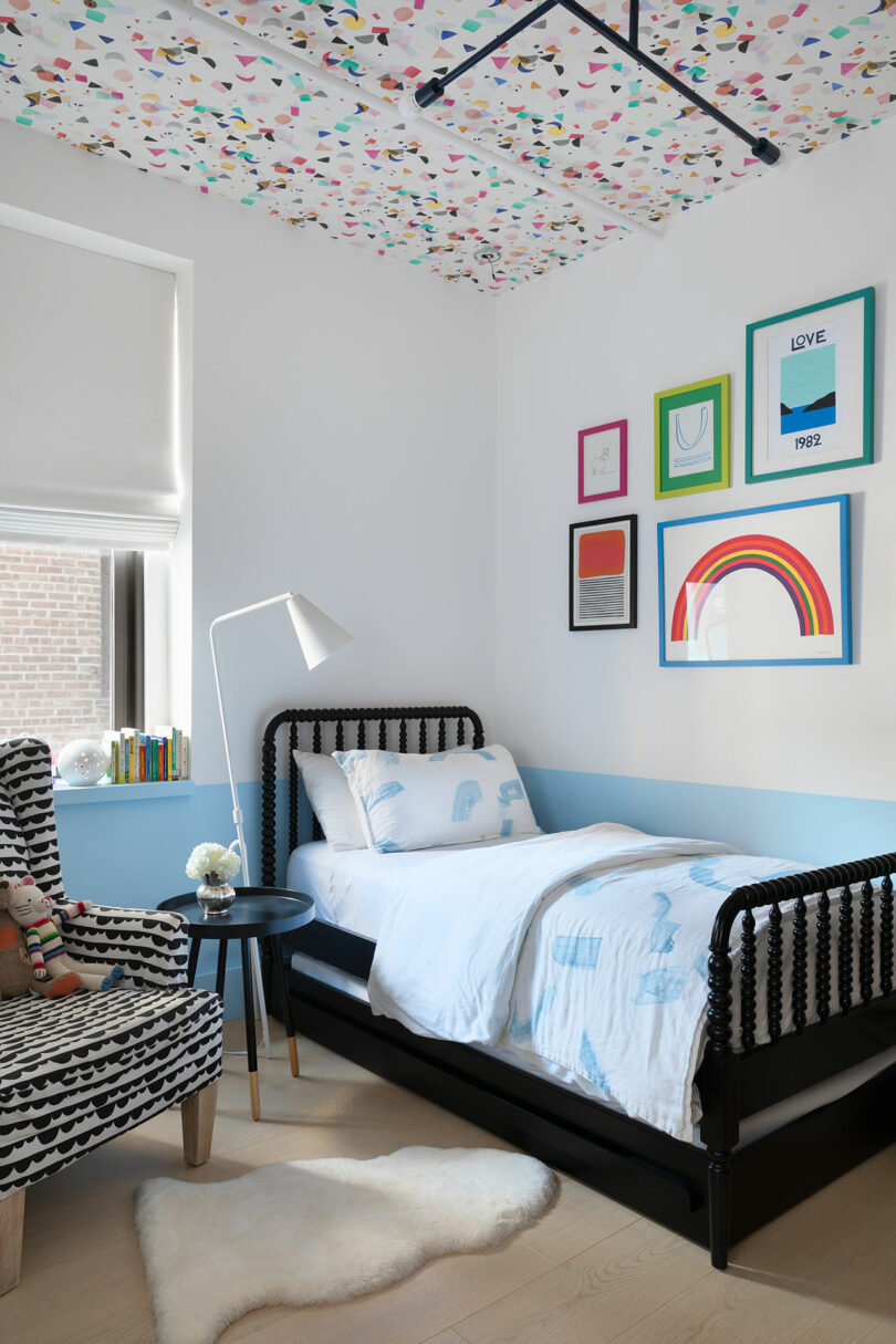 A child's bedroom with a black bed, patterned chair, white floor lamp, colorful ceiling and art, white walls and a shaded window.  The bed has white and blue bedding with animal prints.