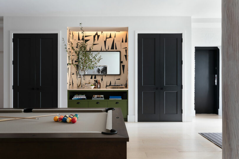A modern room features a pool table with a set of balls and cue stick, flanked by two closed black doors and a niche decorated with a mirror, greenery and abstract wallpaper.