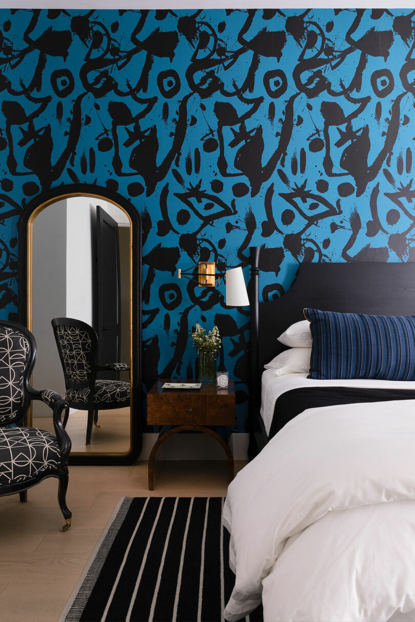 Modern bedroom with black and blue art wallpaper, a white linen bed, a floral wooden nightstand, a black and white striped rug, and a black ornate chair next to a long, gold-framed mirror.