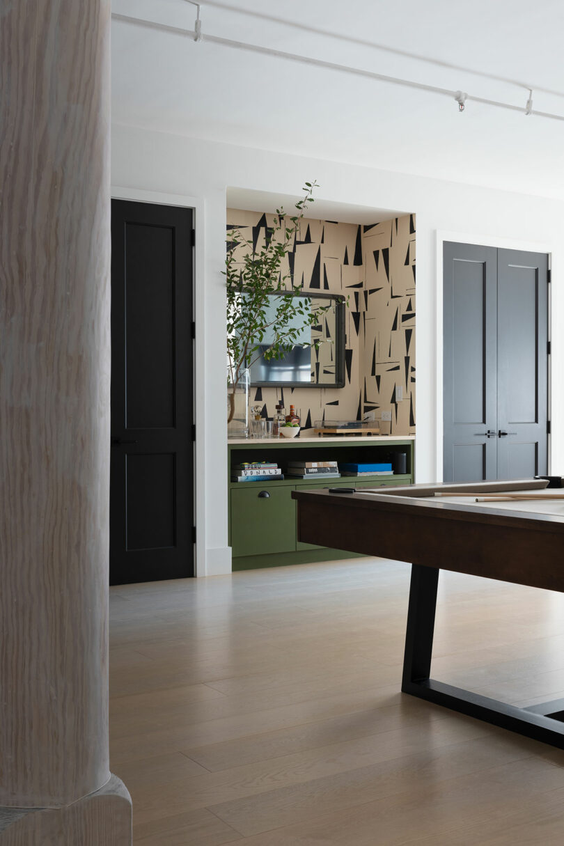 A modern room with a pool table, black cabinets, a green console with books, a TV on a wall with geometric patterns, light hardwood floors and track lighting on the ceiling.