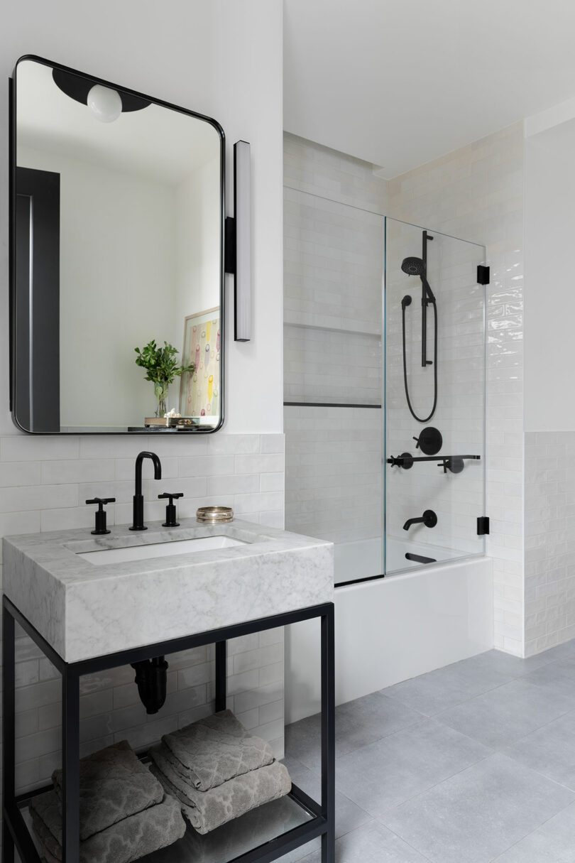 Modern bathroom with a marble-clad vanity unit and black fittings, a floating towel rack below, a glass-enclosed bath with black shower fittings and a large mirror above the sink.