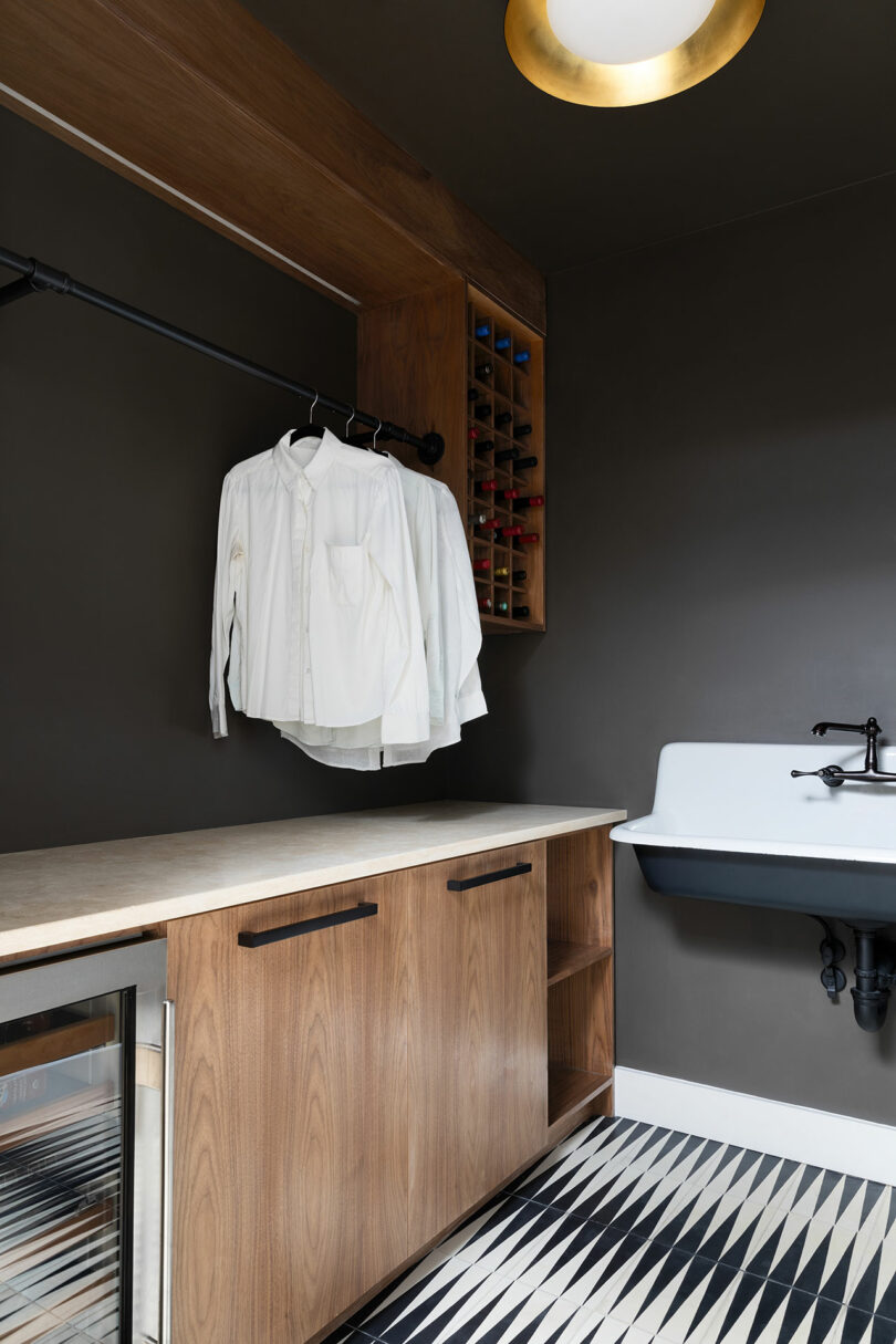 A laundry room with a dark wall, wooden cabinets, a counter, a white sink, a hanging rail with white shirts, a built-in wine rack and patterned floor tiles.