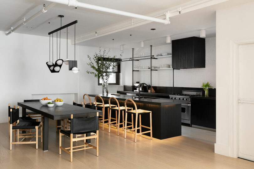 Modern kitchen and dining area with black cabinetry, light wood floors, a long black dining table with chairs, a kitchen island with stools, pendant lights and a potted plant on the counter.