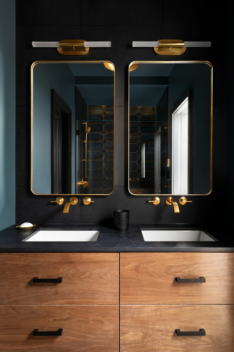 Modern bathroom with two rectangular mirrors, gold fittings and a wooden toilet with two sinks.  Light fixtures are mounted above each mirror.  Black tables contrast with the dark wall behind.